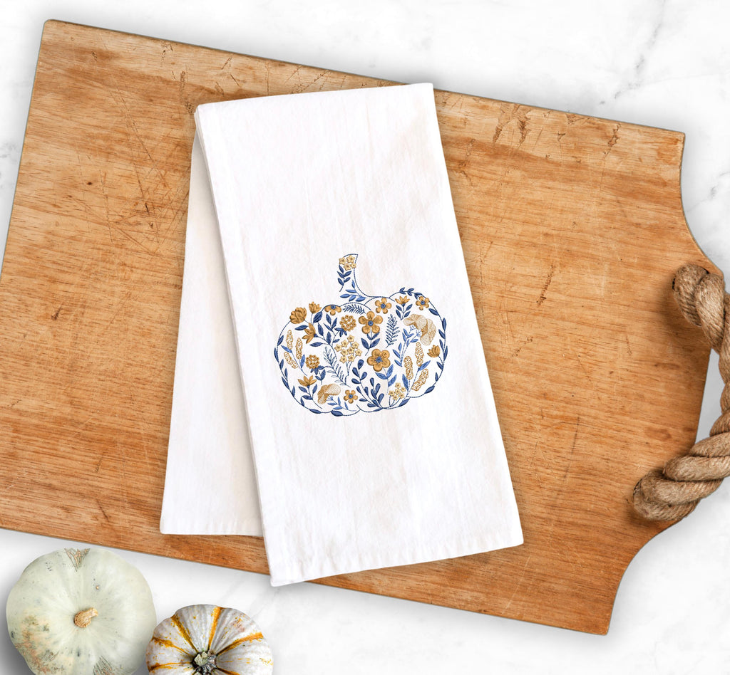Autumnal home decor of a blue and golden orange Embroidered Pumpkin, on top of a cutting board with a rope handle and pumpkins to the lower left  used for Fall Hosting Decor, Thanksgiving Towel, Hostess Gifts, for Your Best Friend.