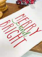 White holiday dish towel with Merry and Bright embroidered on it sitting on a cutting board with cookies and winter berries. Perfect Secret Santa gift for coworker.