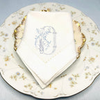 The letter O embroidered on a cotton hemstitch border napkin. The stitching is in blue. It sits upon two antique floral pattern plates on top of a light blue linen tablecloth. These napkins can be personalized with any letter in different colors.