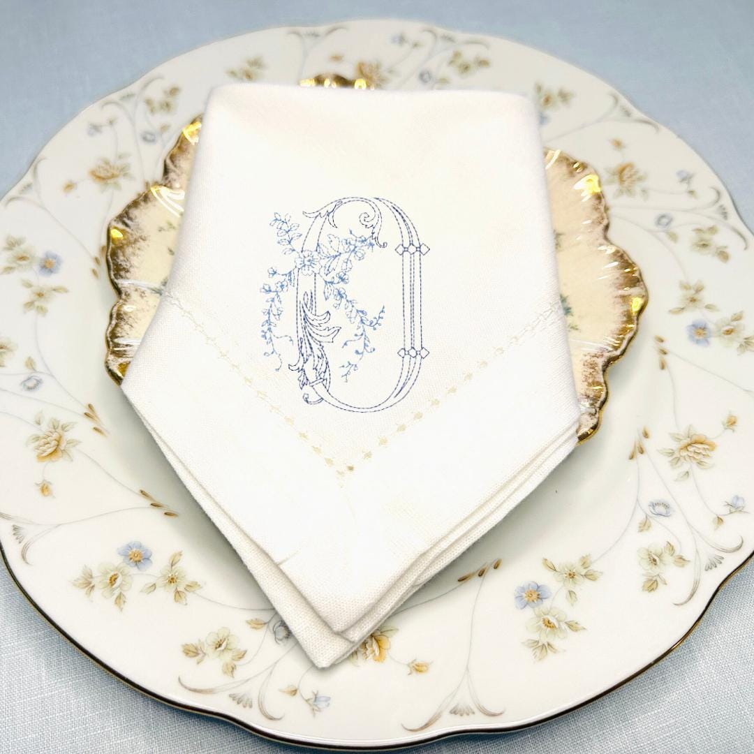The letter O embroidered on a cotton hemstitch border napkin. The stitching is in blue. It sits upon two antique floral pattern plates on top of a light blue linen tablecloth. These napkins can be personalized with any letter in different colors.