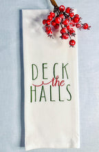 White tea towel set on a light blue linen tablecloth stitched with the words Deck the Halls. There is a berry sprig above the towel.