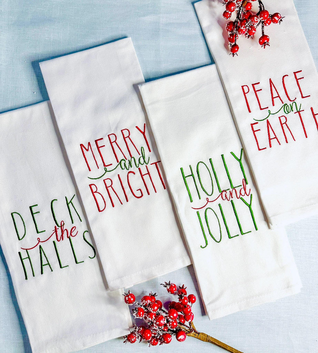 4 White tea towels set on a blue linen tablecloth with red berry branches. This set of towels has four different sayings: Deck the Halls,Merry and Bright, Holly and Jolly, and Peace on Earth. The perfect gift set female best friend.