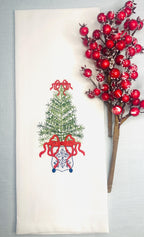 Simple, elegant chinoiserie style Christmas tree stitched on a white tea towel set upon a light blue linen tablecloth with a red berry branch. There is a red bow on top and the bottom of the tree with blue chinoiserie stitching on a white pot.