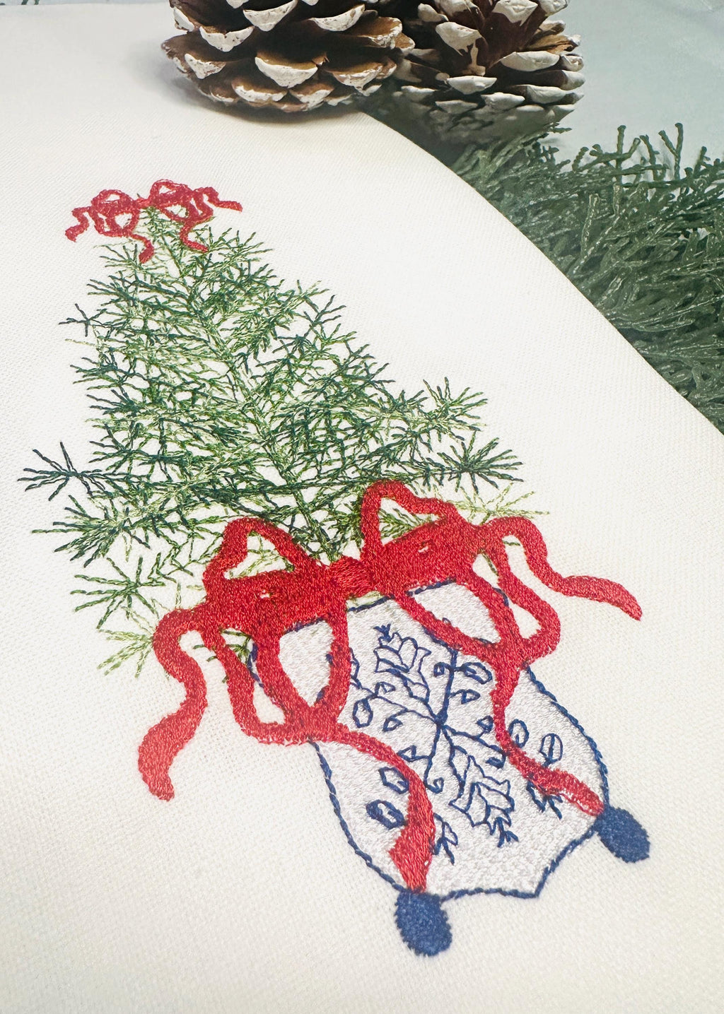 Close up of a simple, elegant chinoiserie style Christmas tree stitched on a white tea towel. There is a red bow on top and the bottom of the tree with blue chinoiserie stitching on a white pot.