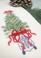 Close up of a simple, elegant chinoiserie style Christmas tree stitched on a white tea towel. There is a red bow on top and the bottom of the tree with blue chinoiserie stitching on a white pot.