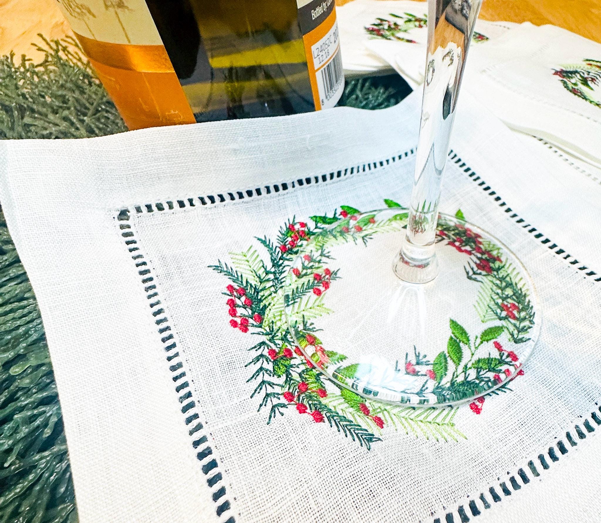 Christmas wreath with different color greens and red berries embroidered on a white cocktail napkin. A wine glass is placed on the napkin next to a bottle of wine on Christmas greens. Perfect gift for Mother in Law Christmas.