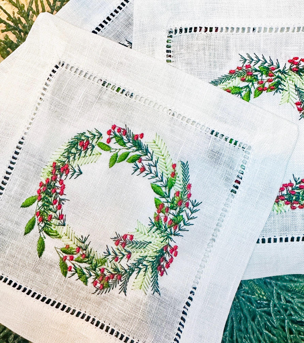Christmas wreath with different color greens and red berries embroidered on a white cocktail napkin. Perfect gift for Mother in Law Christmas. There are three napkins on an evergreen placemat.