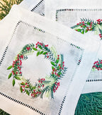 Christmas wreath with different color greens and red berries embroidered on a white cocktail napkin. Perfect gift for Mother in Law Christmas. There are three napkins on an evergreen placemat.