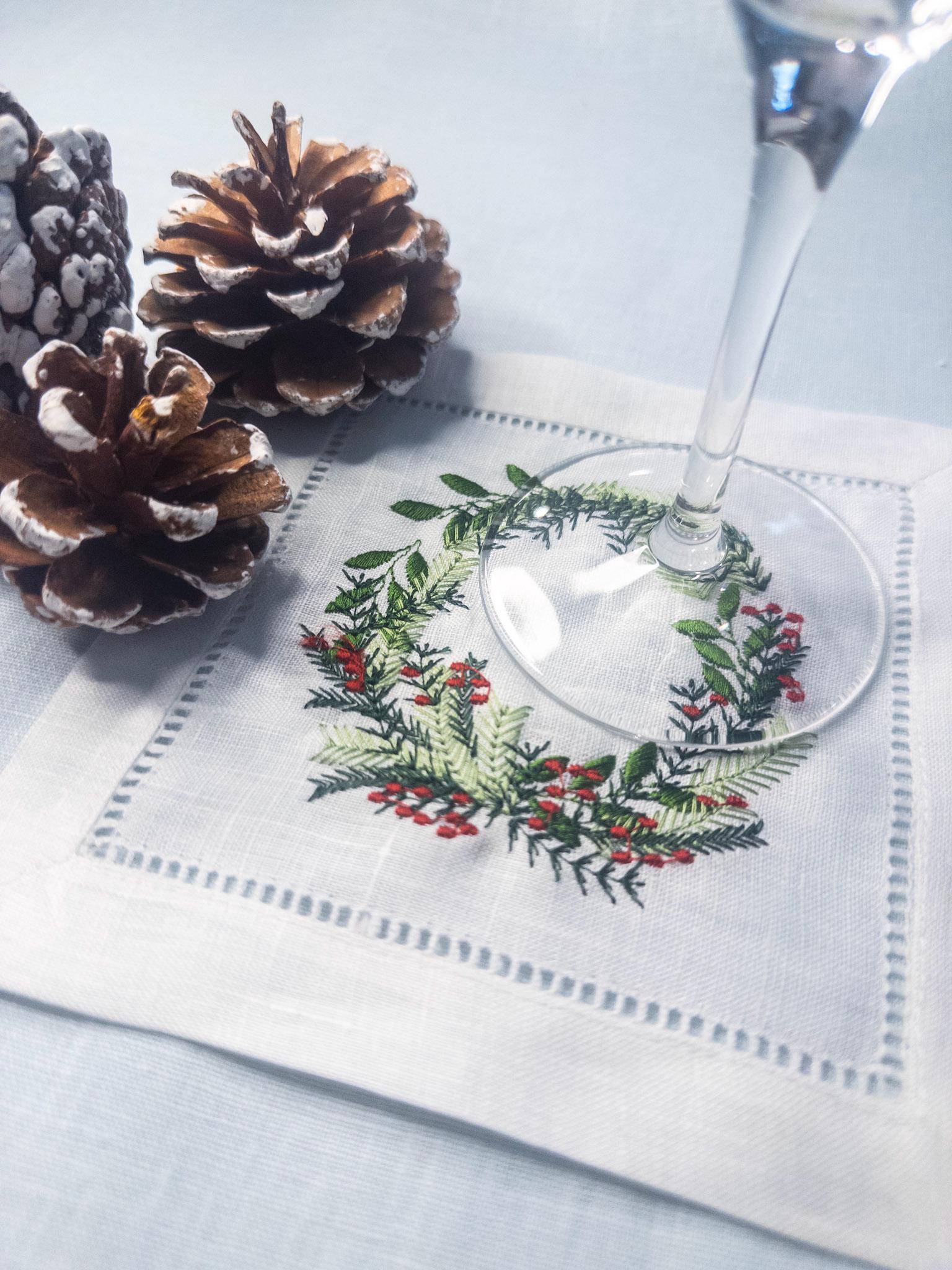 Christmas wreath with different color greens and red berries embroidered on a white cocktail napkin and set on a pale blue linen tablecloth with acorns and a wine glass. Perfect gift for Mother in Law Christmas.