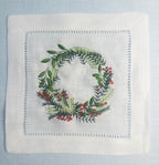A beautiful white linen cocktail napkin with a wreath featuring different color greens and red berries. Perfect Christmas gift for Mother in Law!
