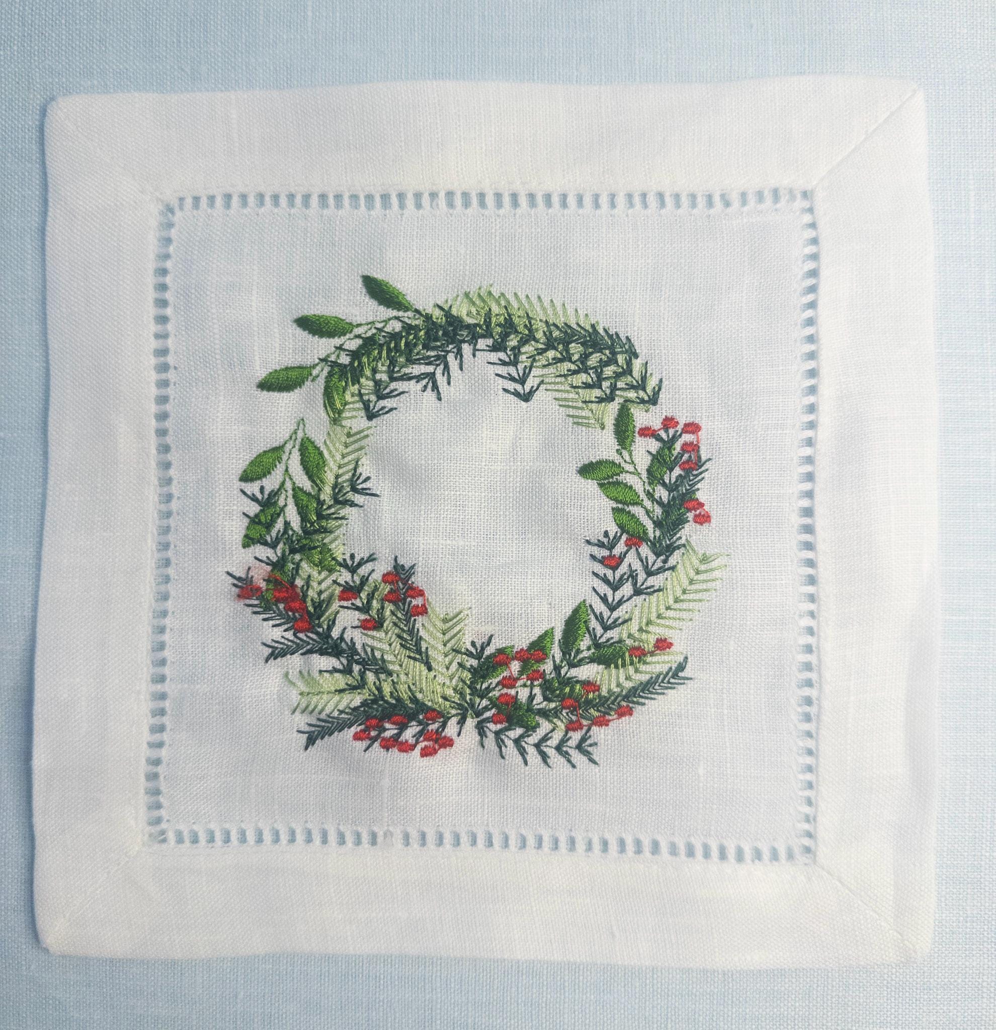 A beautiful white linen cocktail napkin with a wreath featuring different color greens and red berries. Perfect Christmas gift for Mother in Law!