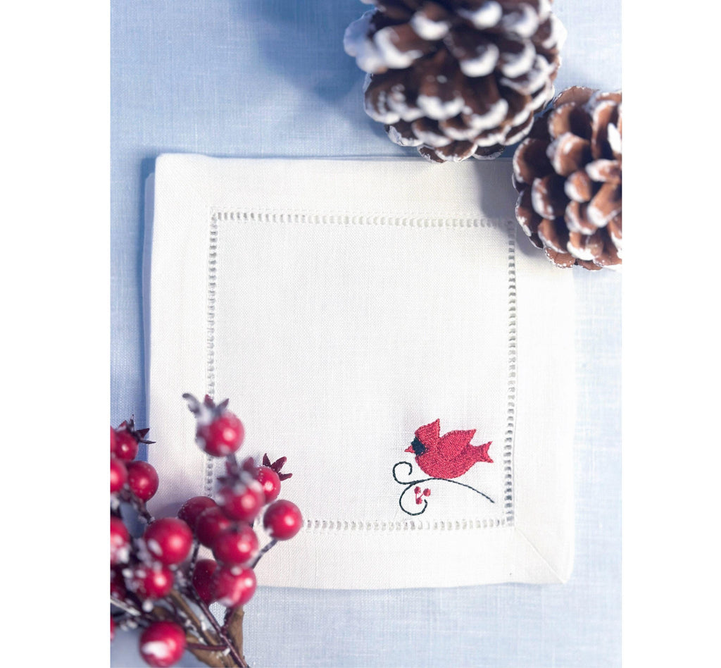 White linen cocktail napkin with a red male cardinal embroidered in the bottom right corner of the hemstitch napkin set on a light blue linen tablecloth with pine cones on top, and Christmas berries on the bottom - for bird lover gift.