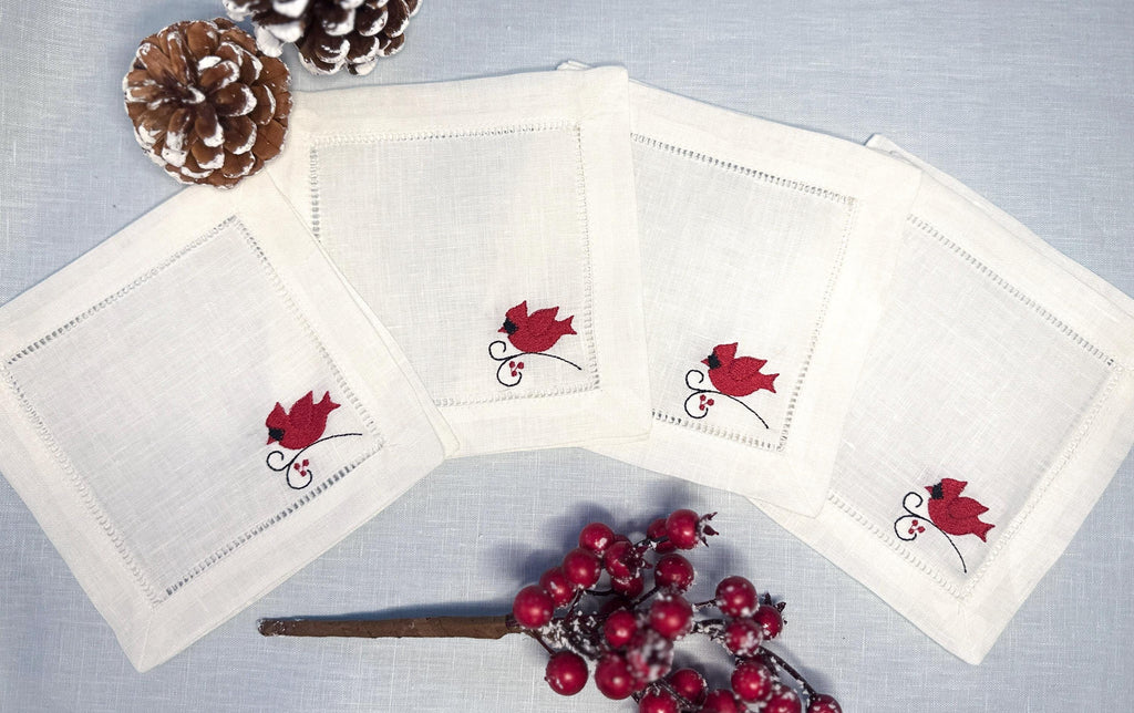 Set of 4 white linen cocktail napkins with a red male cardinal embroidered in the bottom right corner of the hemstitch napkin set on a light blue linen tablecloth with pine cones on top, and Christmas berries on the bottom - for bird lover gift.