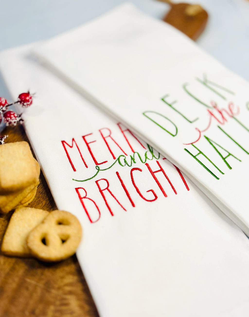 Set of two holiday tea towels one that is embroidered with Merry and Bright and the other with Deck the Halls on a cutting boar with cookies. Perfect coworker Secret Santa gift.