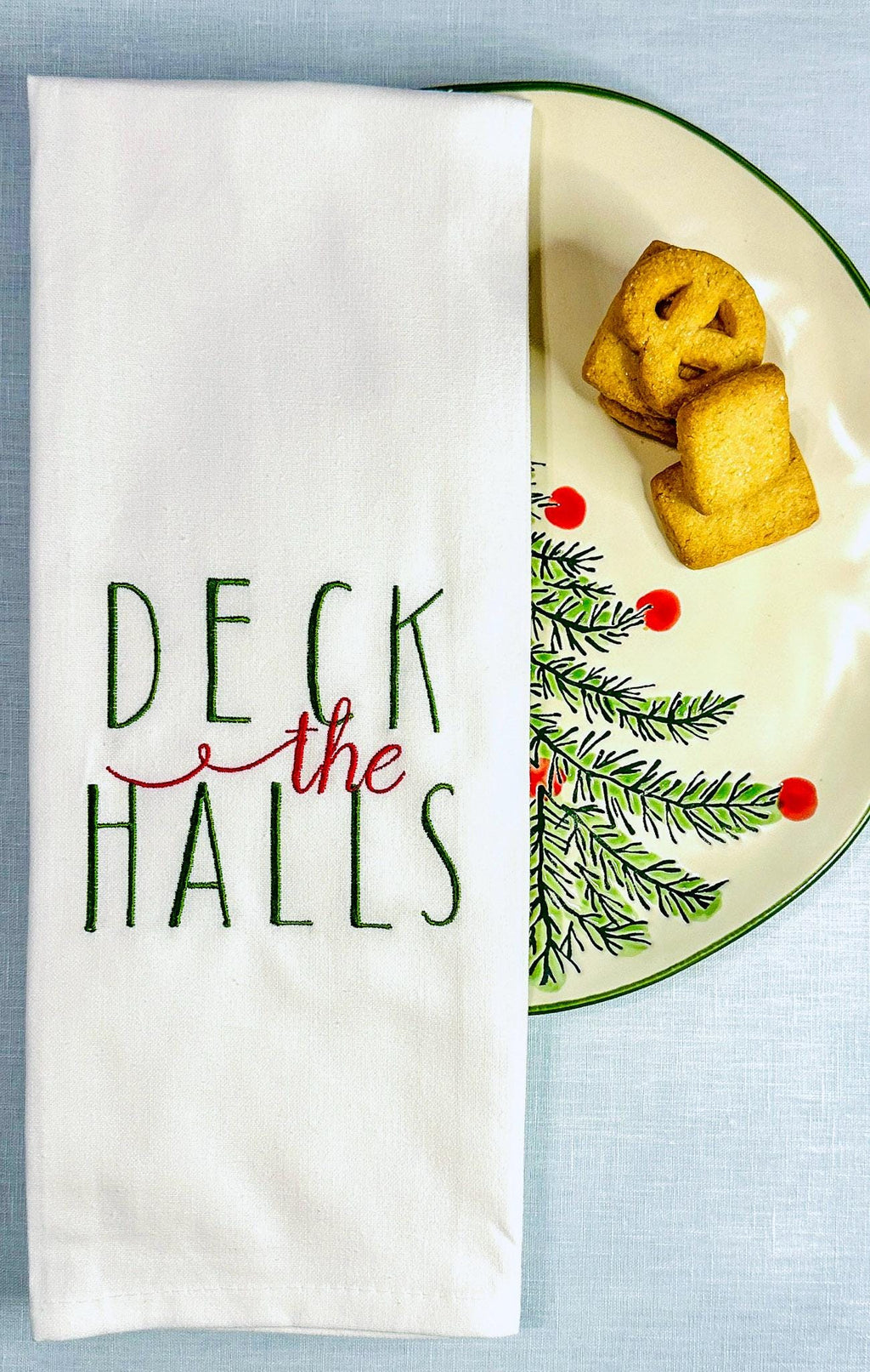 White tea towel with Deck the Halls embroidered in green and red set on a Christmas plate with a holiday tree and cookies.
