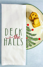 White tea towel with Deck the Halls embroidered in green and red set on a Christmas plate with a holiday tree and cookies.