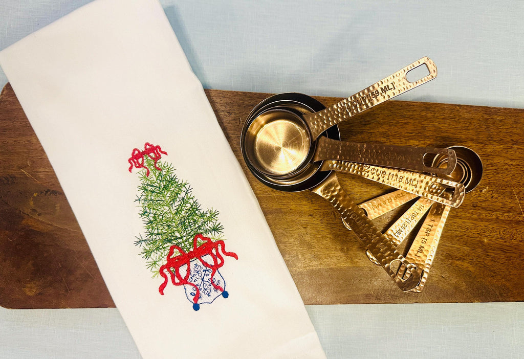 Simple, chinoiserie inspired Christmas tree stitched on a white tea towel set upon a cutting board with copper measuring cup and spoons. There is a red bow on top and the bottom of the tree with blue chinoiserie stitching on a white pot.