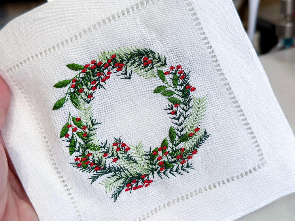 Close up of Christmas wreath with different color greens and red berries embroidered on a white cocktail napkin. Perfect gift for Mother in Law Christmas.