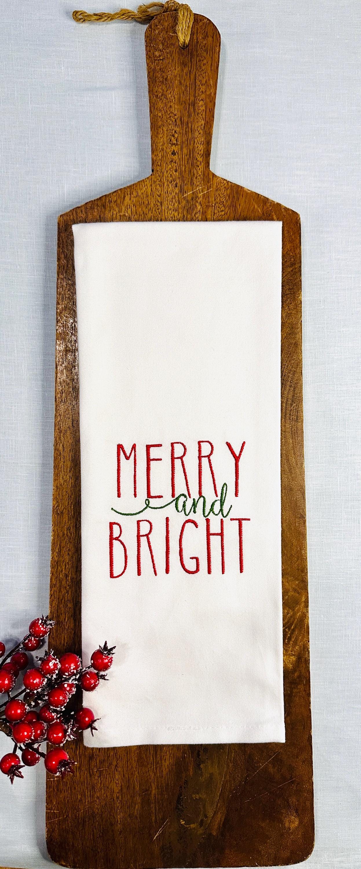 White tea towel on a cutting board on top of a blue linen tablecloth with winter berries. Merry and bright is embroidered on the towel and winter berries as a gift for secret santa coworker.