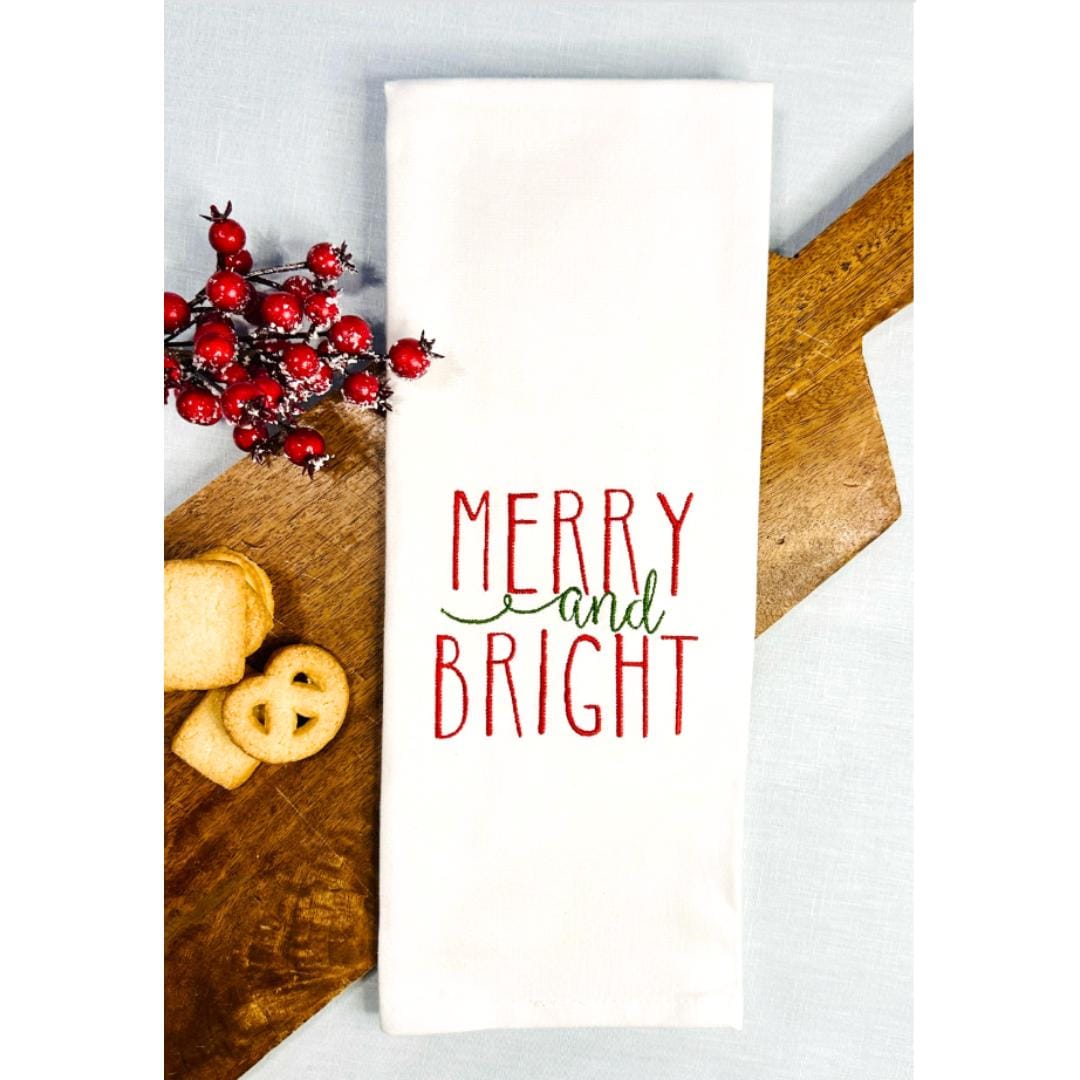 White tea towel on a cutting board with cookies and berry branches embroidered with Merry and Bright as a Secret Santa gift for coworker.
