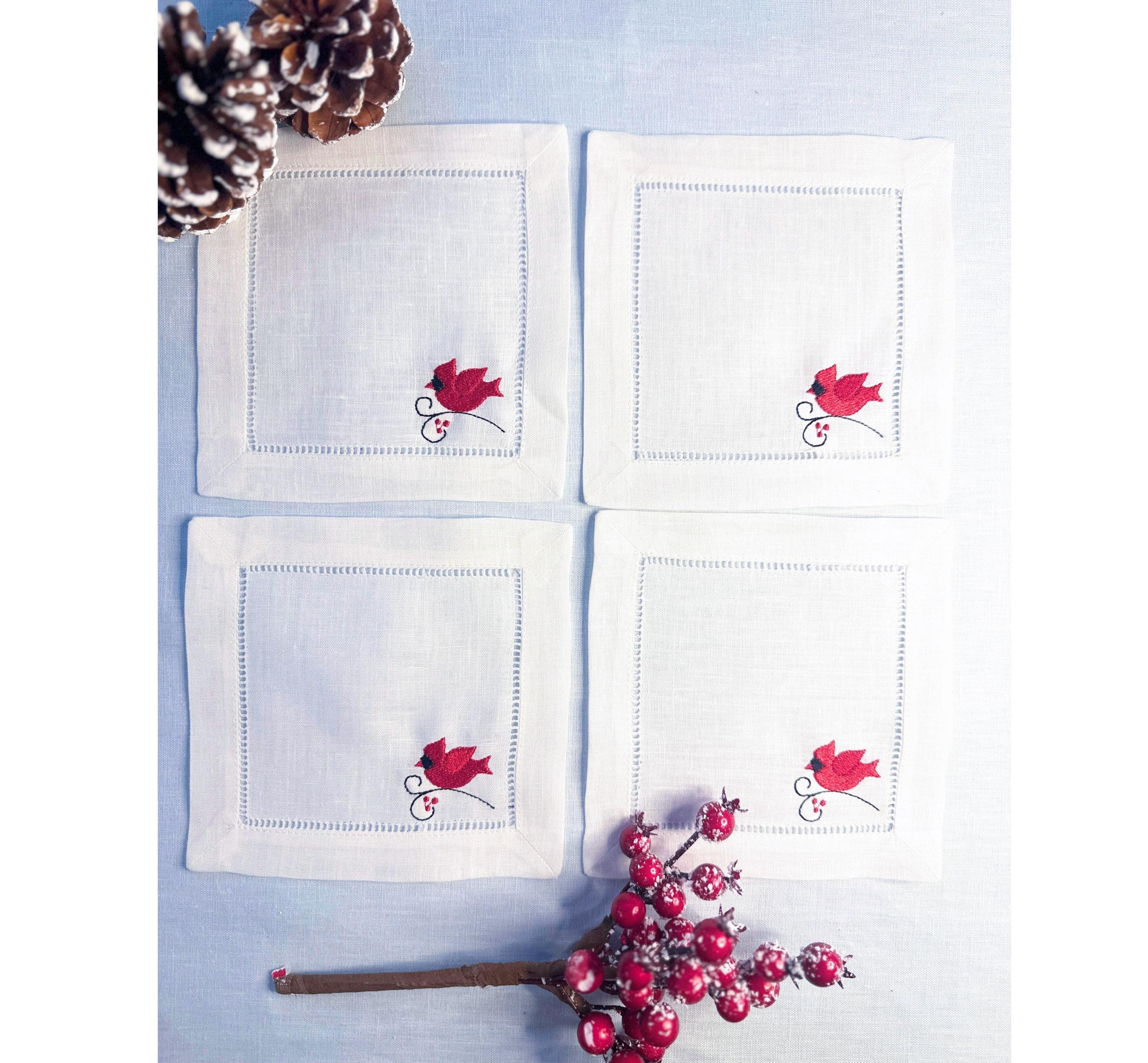 Set of 4 white linen cocktail napkins with a red male cardinal embroidered in the bottom right corner of the hemstitch napkin set on a light blue linen tablecloth with pine cones on top, and Christmas berries on the bottom - for bird lover gift.