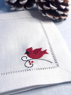 White linen cocktail napkins with a red male cardinal embroidered in the bottom right corner of the hemstitch napkin set on a light blue linen tablecloth with pine cones on top for bird lover or memorial gift.