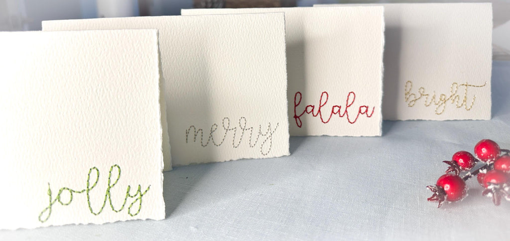 Set of 4 embroidered holiday cards each stitched with a word in a different color: bright (in gold), merry (in silver), jolly (in green), and falala (in red) on a blue linen table cloths with red berries Perfect teacher gift.
