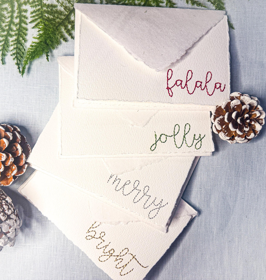 Set of 4 embroidered holiday cards each stitched with a word in a different color: bright (in gold), merry (in silver), jolly (in green), and falala (in red) on a blue linen table cloths with pinecones and greenery. Perfect gift for school secretary.