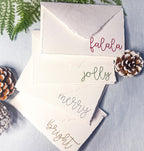 Set of 4 embroidered holiday cards each stitched with a word in a different color: bright (in gold), merry (in silver), jolly (in green), and falala (in red) on a blue linen table cloths with pinecones and greenery. Perfect gift for school secretary.