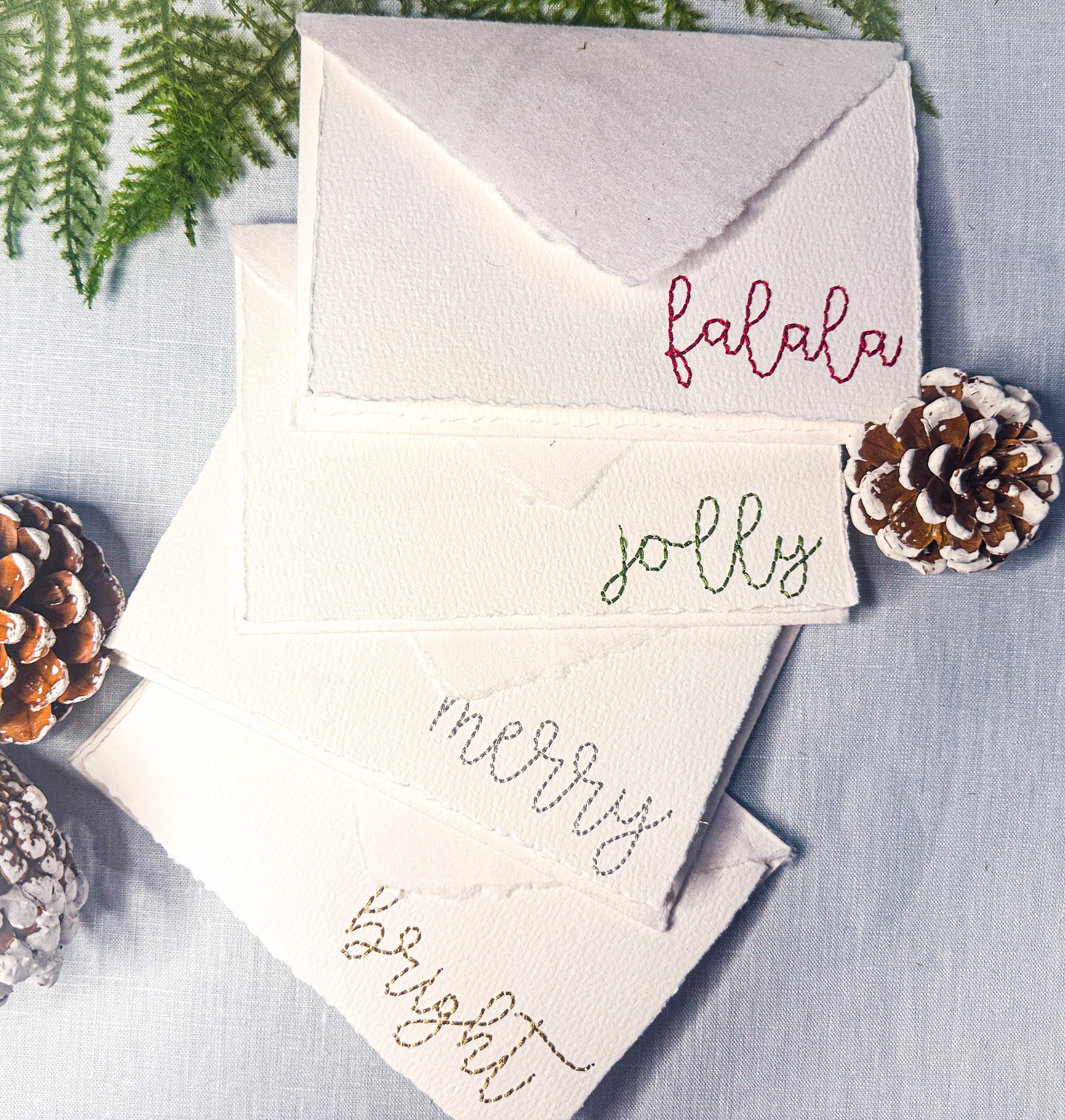 Set of 4 embroidered holiday cards each stitched with a word in a different color: bright (in gold), merry (in silver), jolly (in green), and falala (in red) on a blue linen table cloths with pinecones and greenery. Perfect gift for school secretary.