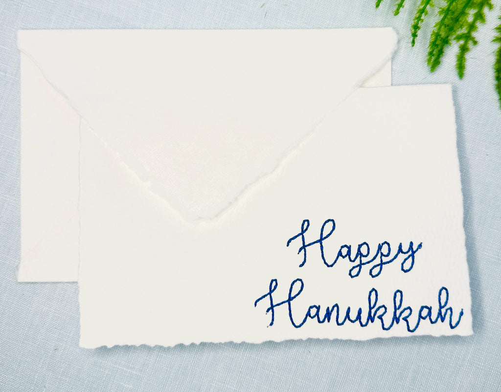 Deckled edge 100 percent handmade cotton greeting cards - set of 4 embroidered in blue with Happy Hanukkah. Cards are bundled with twine and set on a blue linen tablecloth with greenery.