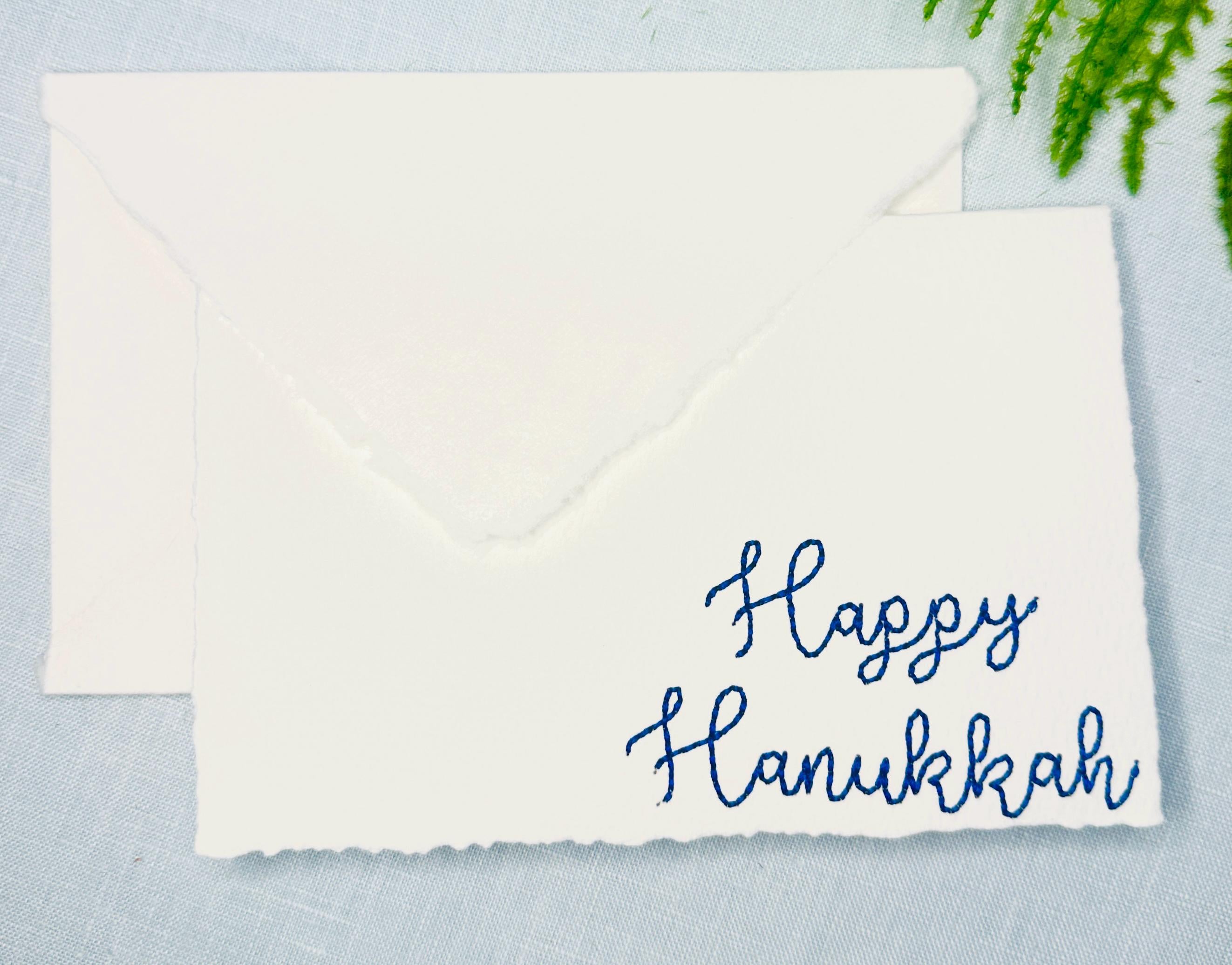 Deckled edge 100 percent handmade cotton greeting cards - set of 4 embroidered in blue with Happy Hanukkah. Cards are bundled with twine and set on a blue linen tablecloth with greenery.