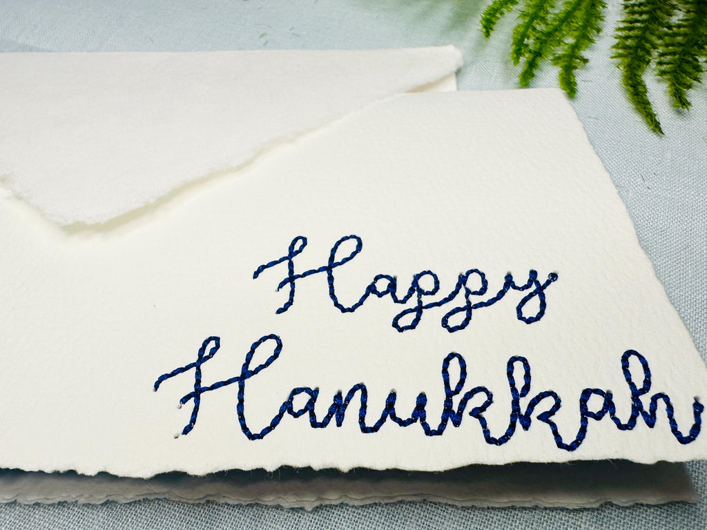 Deckled edge 100 percent handmade cotton greeting card close up embroidered in blue with Happy Hanukkah. Card is set on a blue linen tablecloth with greenery.