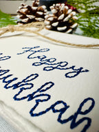 Close up of deckled edge 100 percent handmade cotton greeting card embroidered in blue with Happy Hanukkah. Set on a blue linen tablecloth with two acorn and greenery.