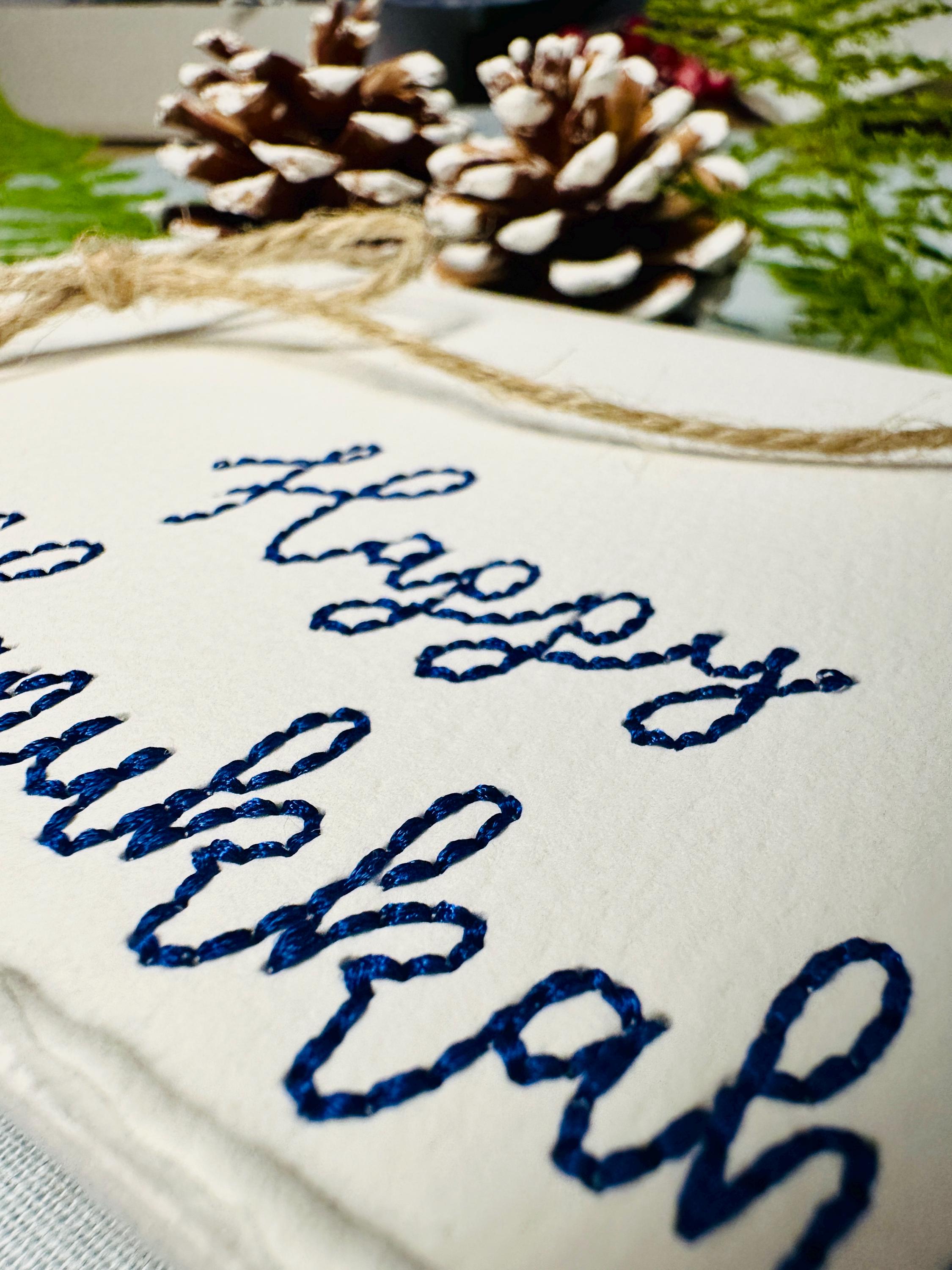 Close up of deckled edge 100 percent handmade cotton greeting card embroidered in blue with Happy Hanukkah. Set on a blue linen tablecloth with two acorn and greenery.