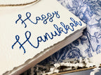 Deckled edge 100 percent handmade cotton greeting cards - set of 4 embroidered in blue with Happy Hanukkah. Cards are bundled with twine and in a box with white crinkle paper and blue and white chinoiserie paper ready to be sent to a customer.