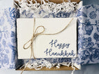 Set of 4 embroidered cards in blue with Happy Hanukkah. Cards are bundled with twine and prepared for gifting set in a white box with white crinkle paper and blue and white chinoiserie tissue.