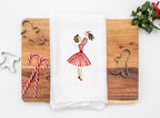 Image of the retro Christmas Decorations 1950s tea towel of mid century modern woman in a red and white striped dress hanging mistletoe. The tea towel is set on a cutting board with cookie cutters, candy canes, and mistletoe.