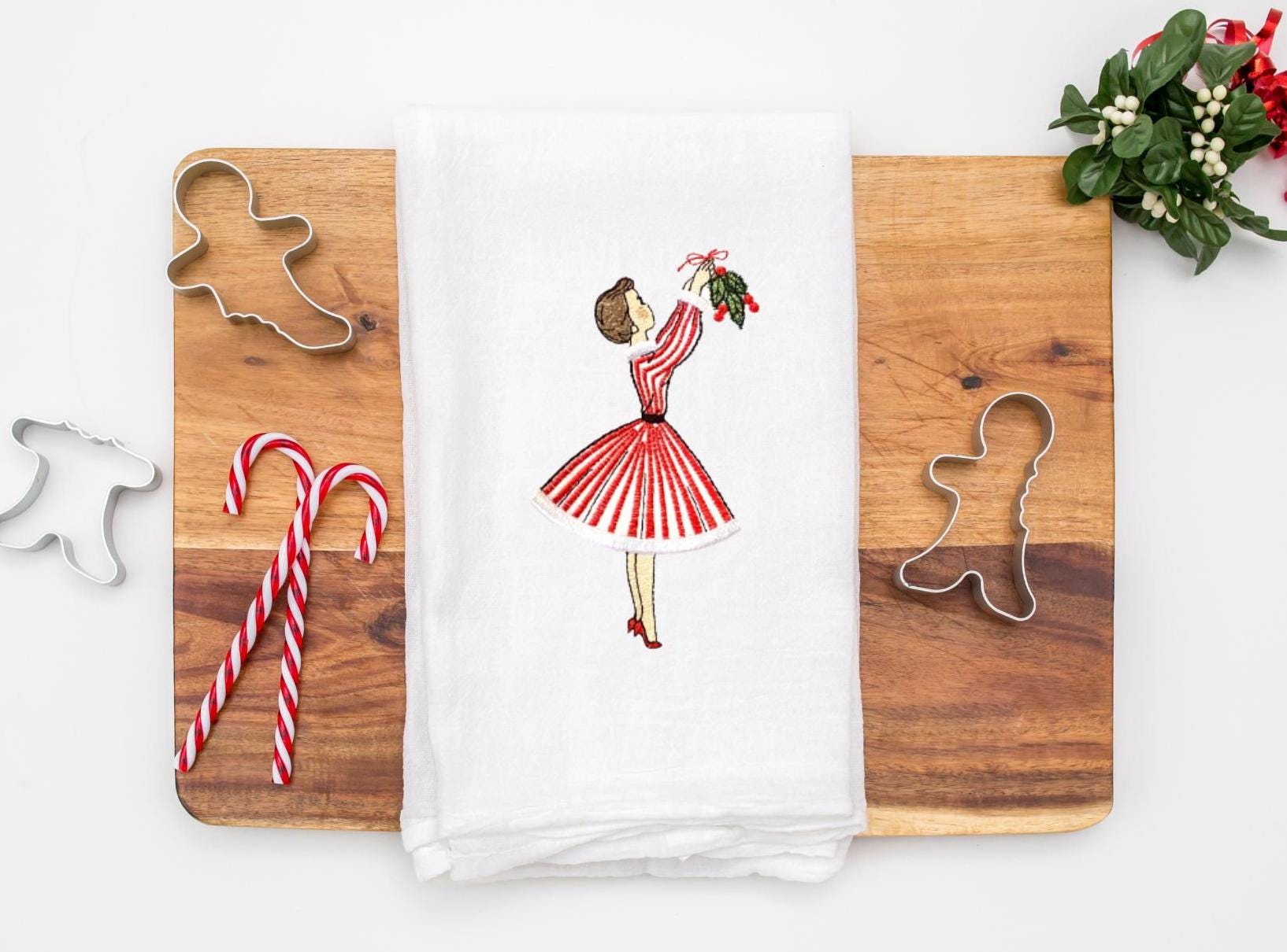 Image of the retro Christmas Decorations 1950s tea towel of mid century modern woman in a red and white striped dress hanging mistletoe. The tea towel is set on a cutting board with cookie cutters, candy canes, and mistletoe.