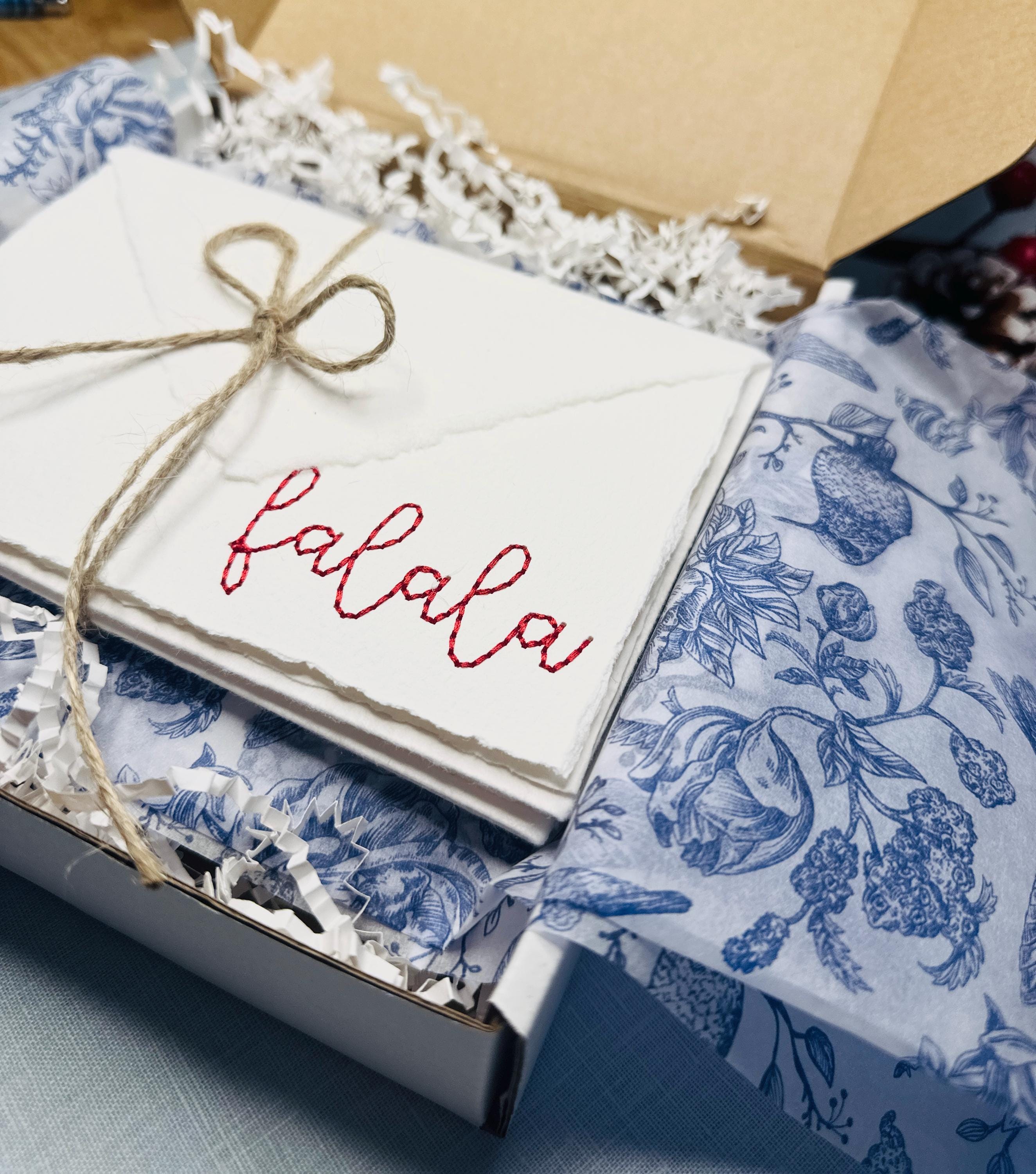 Set of 4 embroidered holiday cards tied in a raffia bow in a white gift bo with white crinkle paper and blue and white chinoiserie tissue paper set on a blue linen table cloth. Perfect gift for teacher.