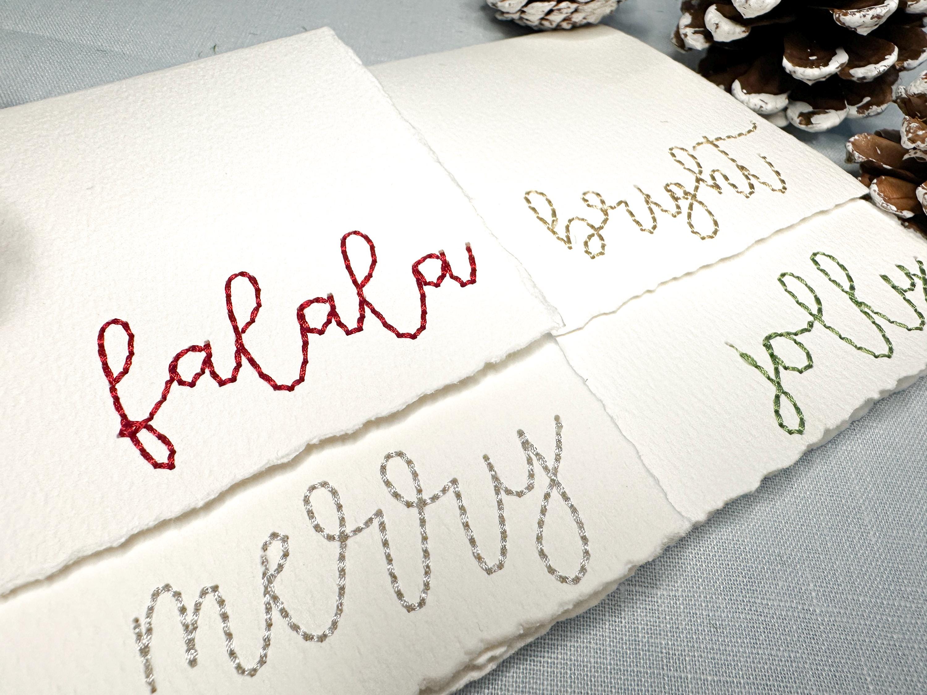 Set of 4 embroidered holiday cards each stitched with a word in a different color: bright (in gold), merry (in silver), jolly (in green), and falala (in red) on a blue linen table cloths with pinecones and greenery. Perfect gift for school secretary.