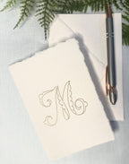 A card from a personalized stationary set of 4 note cards set on a blue linen background highlighting the delicate aesthetic edges of this custom stationary. The embroidery is in gold.