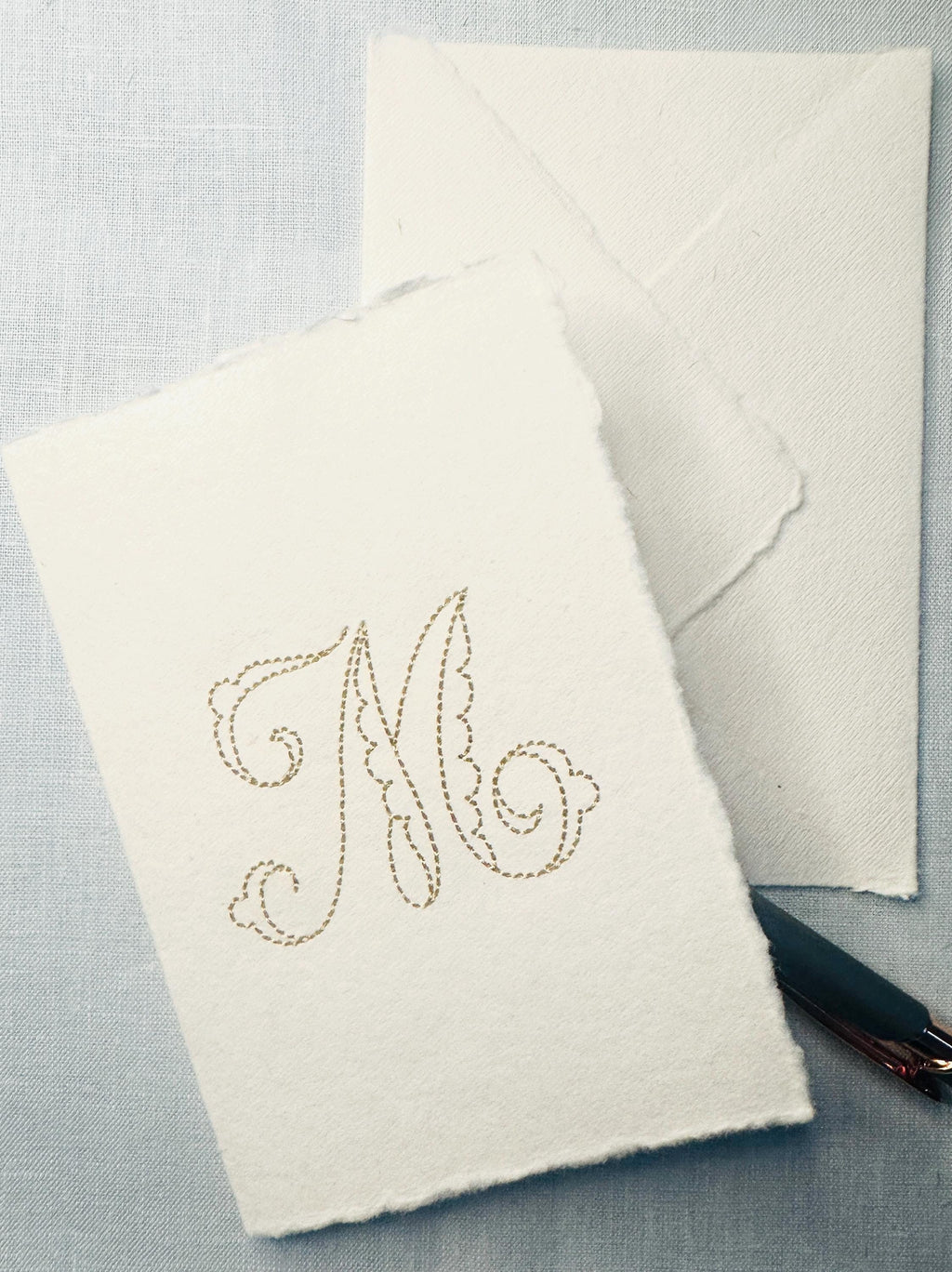 Close up of custom note card embroidered stationery on a blue background with a pen and envelope. The close up shows the deckled edges of the handmade paper used to make the cards.