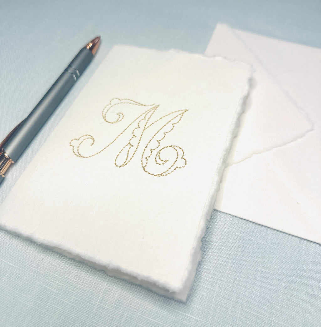Custom note card embellished with an embroidered M as an example of personalized stationary. Set on a blue linen background with a pen to the left hand side and an envelope.