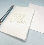 Custom note card embellished with an embroidered M as an example of personalized stationary. Set on a blue linen background with a pen to the left hand side and an envelope.