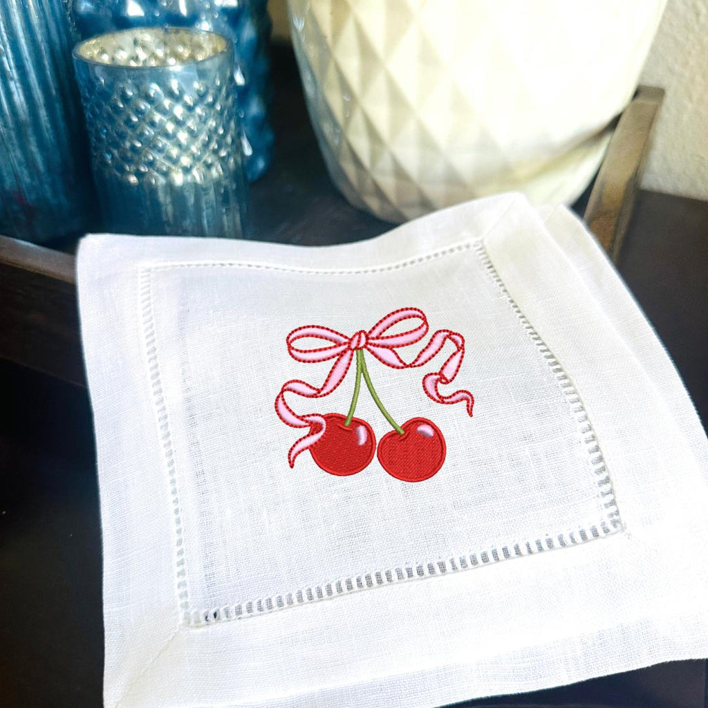 Set of 4 cherry embroidered linen cocktail napkins gift idea for 50th birthday gift for women. Two cherries on stem with a pink bow embroidered on a cocktail napkin and set on a bar with votive holders.