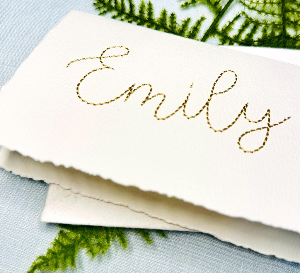 The name Emily is stitched in gold on a 100% cotton handmade paper card. The card is on top of its&#39; matching envelope. They are on a light blue linen tablecloth with a fern.