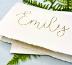 The name Emily is stitched in gold on a 100% cotton handmade paper card. The card is on top of its&#39; matching envelope. They are on a light blue linen tablecloth with a fern.