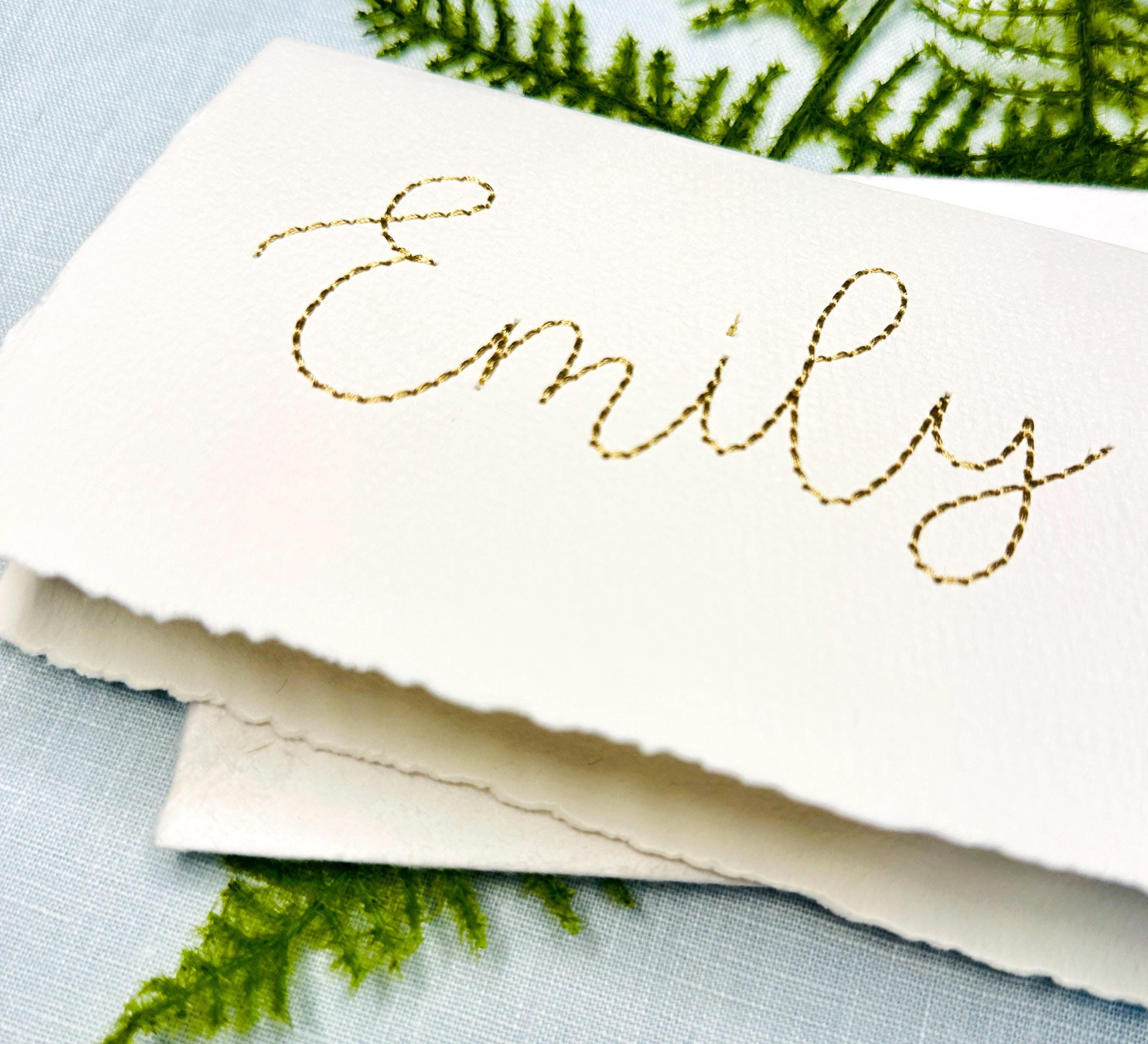 The name Emily is stitched in gold on a 100% cotton handmade paper card. The card is on top of its&#39; matching envelope. They are on a light blue linen tablecloth with a fern.