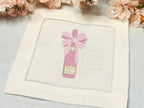 Beautifully embroidered pink champagne bottle with bow stitched onto a white linen cocktail napkin set on a light blue linen tablecloth with cherry blossoms at the top.